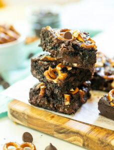 a stack of three brownies with caramel and pretzels on a cutting board