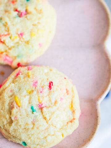 Two cake batter cookies with rainbow sprinkles on a pink platter with scalloped edges.